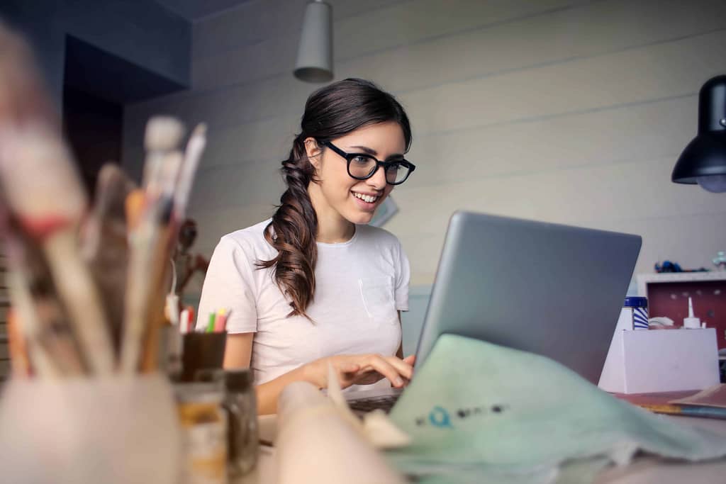 uśmiechnięta, młoda kobieta w okularach siedząca w biurze pisze list motywacyjny po angielsku na komputerze