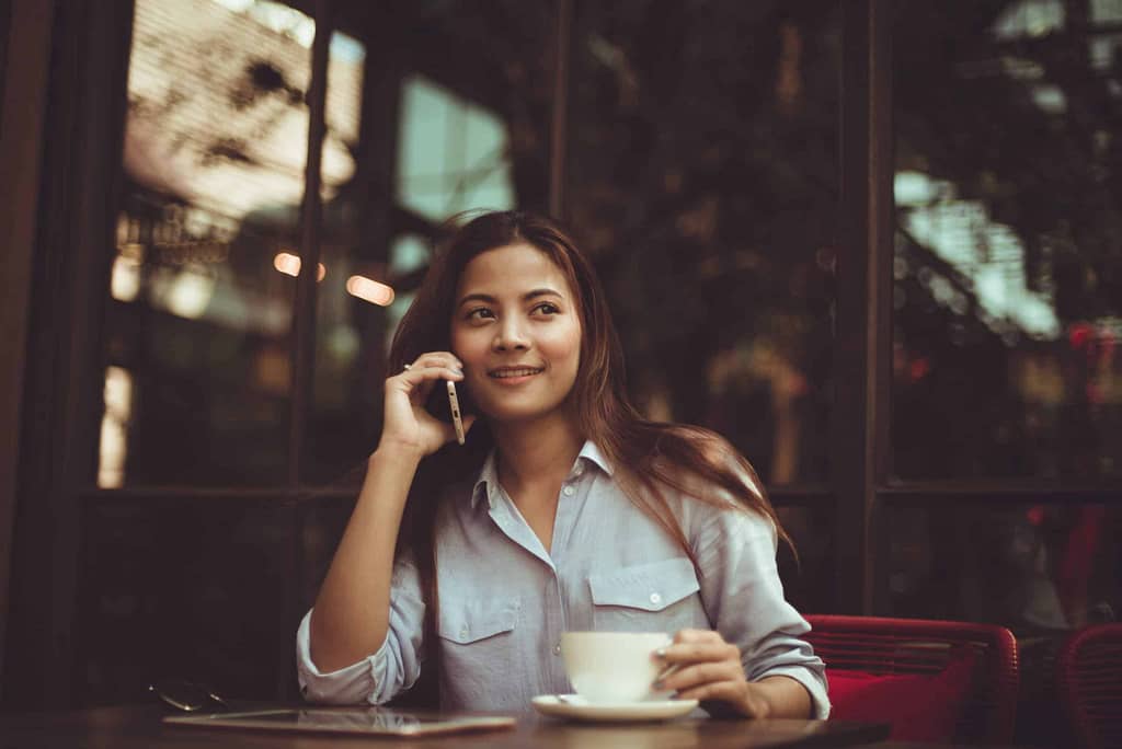 kobieta siedząca w biurze z filiżanką kawy. rozmawia przez telefon po angielsku i uśmiecha się do rozmówcy